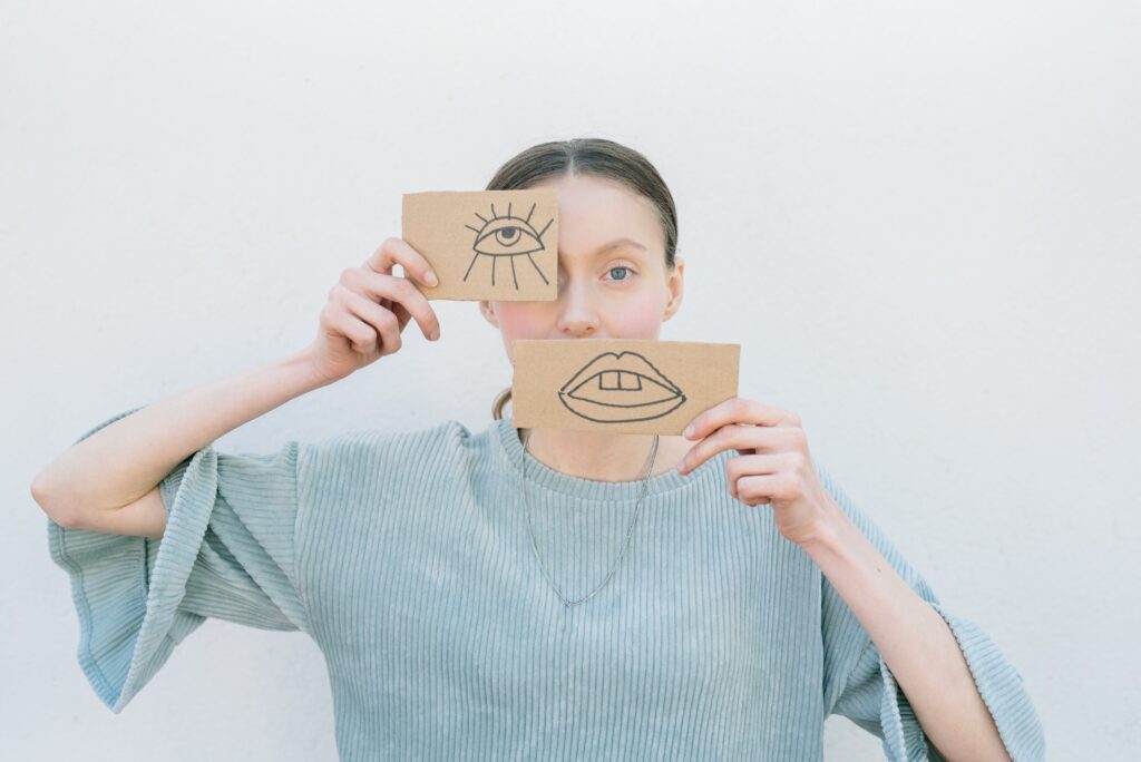 women face covering one eye and mouth with eye and mouth drawn on card board